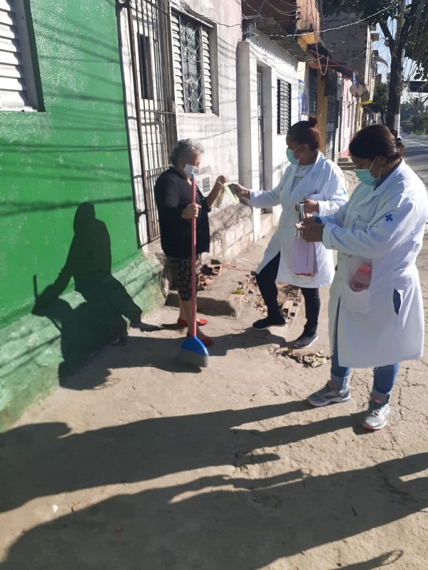 #PraCegoVer: Na imagem, fotografia de duas profissionais da saúde distribuindo máscaras. Elas estão com avental branco.